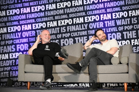 Vincent D'Onofrio and Charlie Cox sit on the main stage on the gray leather couch. Both sitting and smiling at the moderator (out of frame), legs crossed, with their left hands touch the backs of their respective heads