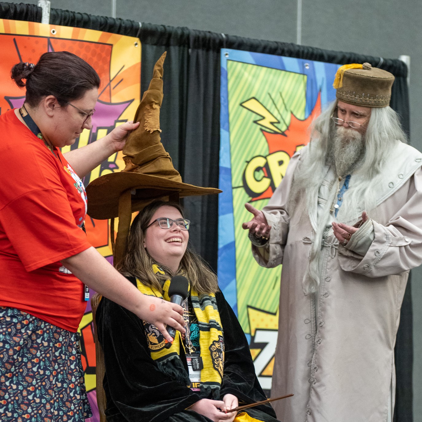 Woman in red dress placing the sorting hat on a girl in a robe an Hufflepuff scarf. Next to them is Dumbledore.