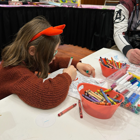 A young girl, wearing an orange sweater, and red hair bow, sits at a white table colouring an image with a green marker. Around her there are various colours of crayons and markers, and to her right, a parent dressed in the Avenger's Endgame suit colours with her.