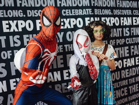Three individuals pose in front of a backdrop filled with the words "FANDOM," "EXPO," "CELEBRATE," and other related phrases. On the left, someone in a detailed Spider-Man costume strikes an action pose, showcasing the iconic red and blue outfit with white web patterns. In the center, a person wears a Spider-Gwen mask and white hoodie, holding up a hand to display their red web-patterned glove. On the right, a young girl with curly hair and green-framed glasses stands confidently. She wears a colorful floral dress and holds a brown handbag. The group seems to be enjoying a pop culture or fandom event.