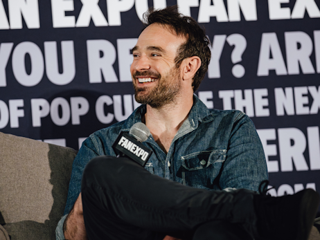Charlie Cox sitting on a couch on stage, holding a microphone and smiling.