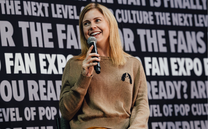 Rose McIver with blonde hair and a bright smile speaks into a microphone. She is wearing a beige sweater with a black silhouette of an image on it. She sits in front of a backdrop with repeated fan expo text, suggesting she's part of a panel or event.