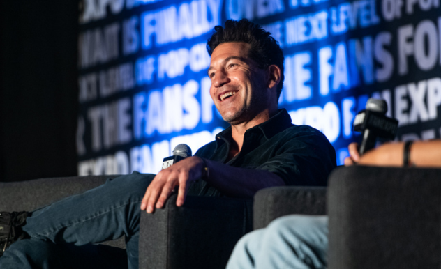The image features Jon Bernthal comfortably seated on a couch, engaging with an audience during a panel discussion at a FAN EXPO event. He holds a microphone in one hand, his relaxed posture and genuine smile conveying a sense of ease and enjoyment. He's dressed casually in a dark shirt and jeans, suggesting a laid-back approach to the event.