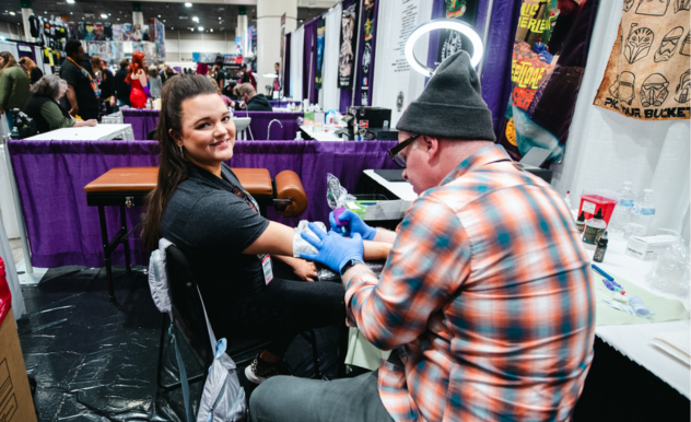 The image depicts a tattoo artist at work, applying a tattoo to the forearm of a smiling woman. They are in an indoor setting that appears to be a convention or expo, indicated by the booths and banners in the background, as well as other people milling about. The artist, wearing gloves for hygiene, is focused on his work, while the woman, comfortably seated, looks directly at the camera. The atmosphere seems busy yet casual, with a mix of professional engagement and personal enjoyment.