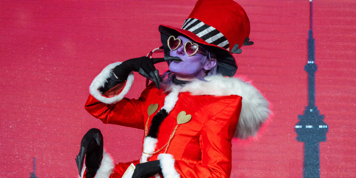A cosplayer painted purple blows a kiss to the audience. Dressed as Valentino from Hazbin Hotel, tehy are sporting extra arms, and an all red suit.