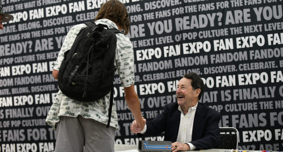 Peter Cullen shakes hands with an eager fan. He is signing an Optimus Prime action figure for them with a huge smile.