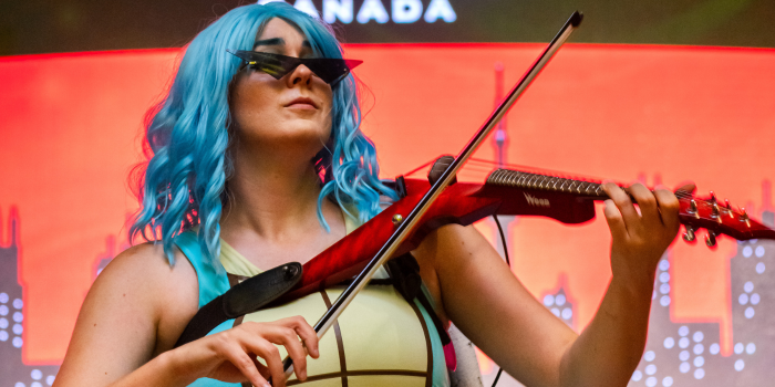 Dressed as one of the Squirtle Squad, Brigit Fiddle treats the audience to a performance at competition half time. She is wearing black angular sunglasses and playing a red v-shaped fiddle. 