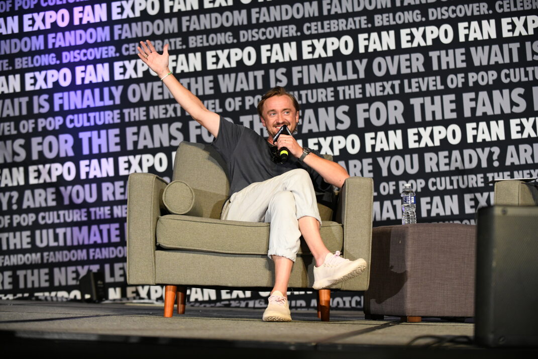Tom Felton greets a large and excited audience. He smiles a large smile and holds his left hand up in excitement as he begins his panel on the main stage