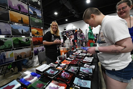 fans browse a selection of handmade coasters and trays, and art prints. The vendor catches the camera and smiles at the photographer