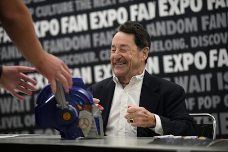 Peter Cullen smiles as a fan places an Optimus Prime helmet on the table to be signed. Peter uncaps a red sharpie to sign the helmet.
