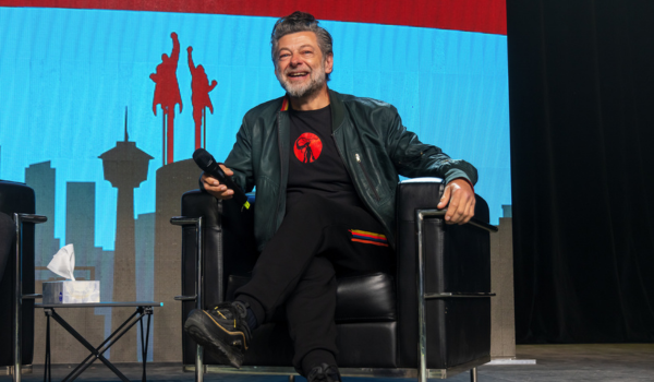 Andy is sitting in a black chair on stage. He is wearing a black leather jacket with a black and red t-shirt underneath.