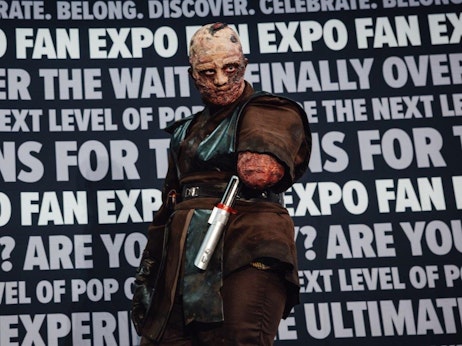 A character with detailed makeup to resemble a scarred and battered face stands in front of a bold backdrop filled with phrases like "FAN EXPO," "CELEBRATE," "DISCOVER," and other event-related words. The character wears a dark leather outfit, and a cylindrical prop, resembling a futuristic weapon, is attached to their belt. The overall ambiance suggests a scene from a pop culture or fandom convention, with the character embodying a menacing presence.