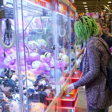 A fan with green dreadlocks, wearing a grey sweater, plays an orange claw machine full of purple stuffed Totoros