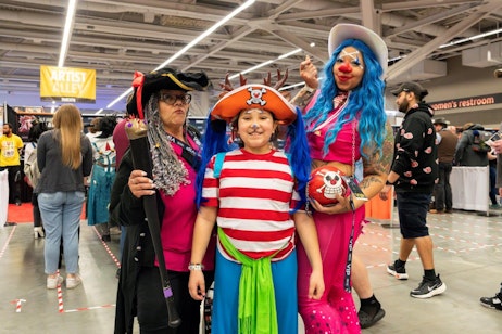 The image captures a joyful family moment at a fan convention with three people dressed in vibrant and playful pirate-themed costumes. On the left, a woman is wearing a traditional pirate hat and carrying a wooden sword, displaying a serious expression. The child in the center smiles widely, dressed in a colorful striped outfit, embodying a more whimsical pirate look with a bright red hat featuring a skull and crossbones. On the right, another woman sports bright blue hair and a vivid pink outfit, exuding a cheerful demeanor as she waves a small pirate flag. The backdrop of the convention center includes a sign for "Artist Alley," hinting at the creative and bustling environment of the event. This family showcases their shared enthusiasm for cosplay and the fun of engaging in character roles together.