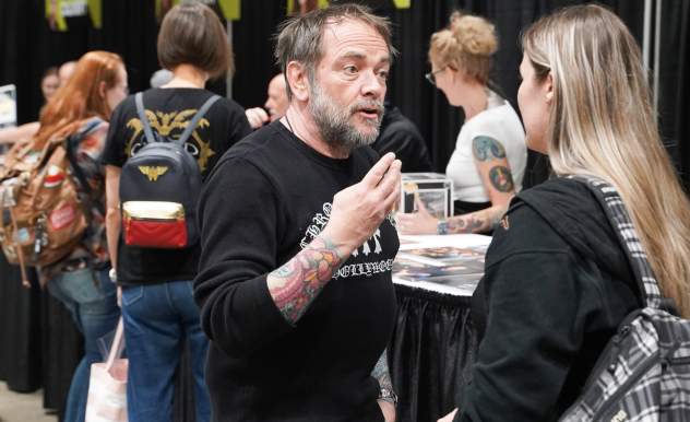 Mark Sheppard has a conversation with a fan, her back faces the camera. He is speaking with his hands and they are raised as he speaks.