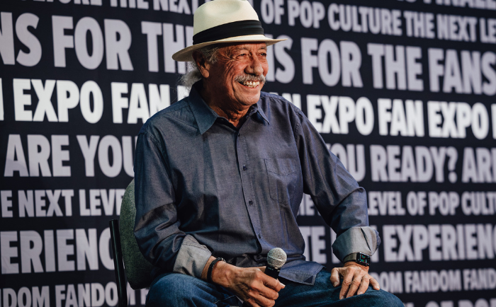 Edward James Olmos with a warm smile, wearing a blue shirt, white hat, and wristwatch sits comfortably with a microphone beside him. He's in front of a backdrop with repetitive fan expo text, indicating his participation in an event or panel discussion.