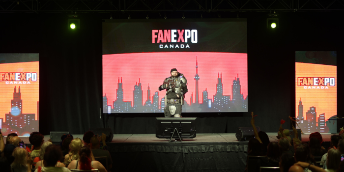 The audience watches and cheers as an armoured individual takes the stage. Blaster facing forward, they strike a pose for the crowd.