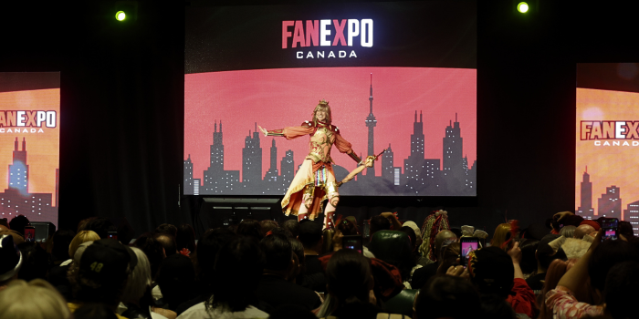 An armoured princess walks the stage, posing with their arms extended. The audience as taking photos as she smiles.