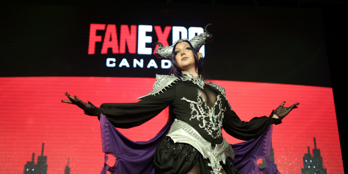 Dressed in black, purple, and grey, a cosplayer poses as a tiefling like creature, arms open and extended showing the attached cape. 