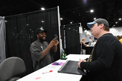 Shameik Moore checks a piece of art on a fan's phone with a huge smile while the fan explains.