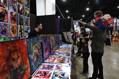 A father points out a piece of art to his two young children. One, dressed as Spider-man, on his shoulders, the other is standing in front of him, dressed as Miles Morales.