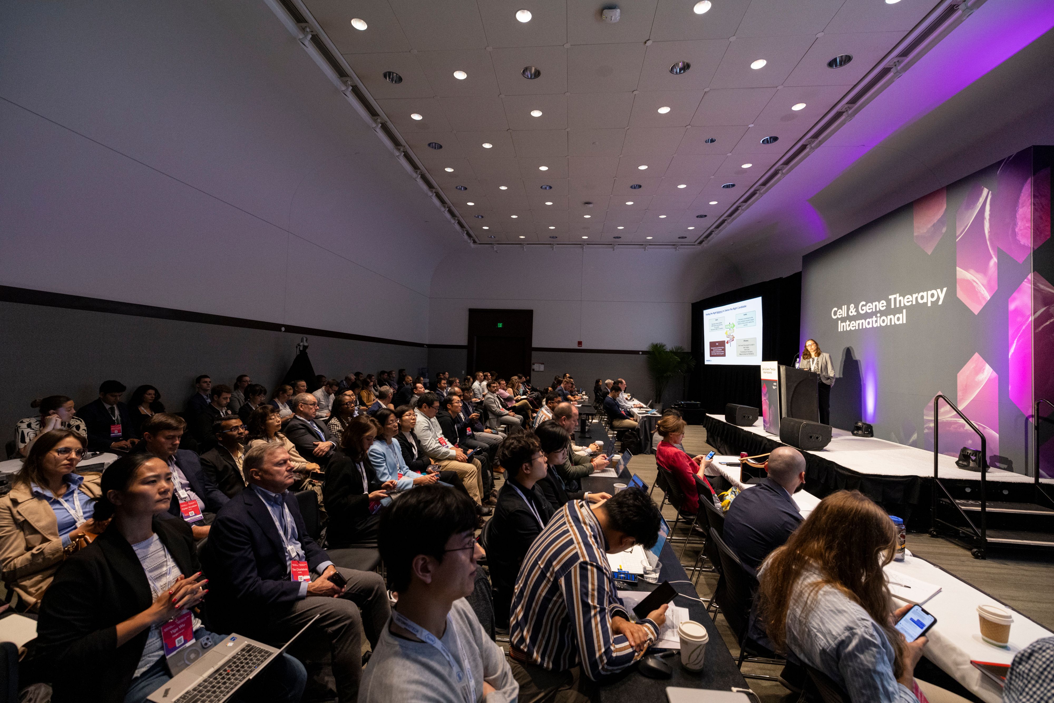 Biopharma attendees sit watching a speaker on stage in a room at Cell & Gene Therapy International.
