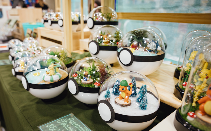 A display of spherical terrariums designed to resemble Pokémon balls, each containing various Pokémon figures set in miniature dioramas. The dioramas feature different scenes, from snow-covered landscapes with sparkling trees to grassy settings with mushrooms. In the forefront, a terrarium showcases an orange Pokémon surrounded by frosty trees. A sign on the table reads "Pokéball Terrarium $30" indicating they are for sale. The background hints at a convention or market setting with other merchandise on display.