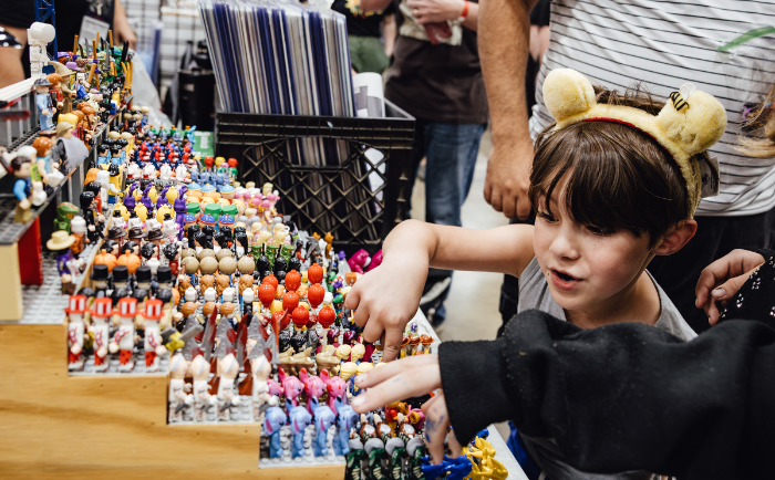 A young boy with bear ear headband excitedly reaches out to touch a display of colorful mini figurines at a convention booth. He has a look of fascination and curiosity on her face. Behind the girl, other attendees and merchandise can be seen, suggesting a lively convention environment. The figurines are meticulously organized, showcasing a variety of characters and designs.
