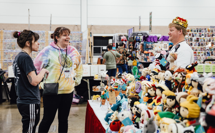Two young women laugh and converse with a vendor wearing a crown at a convention booth. The booth showcases an array of plush toys, figurines, and other merchandise neatly arranged on tables. The atmosphere appears friendly and engaging, with the backdrop revealing other stalls and attendees browsing in a large convention hall. The young woman on the left wears a black top with an "adidas" logo and the other dons a vibrant tie-dye hoodie. The vendor seems animated in conversation, adding to the jovial mood of the scene.