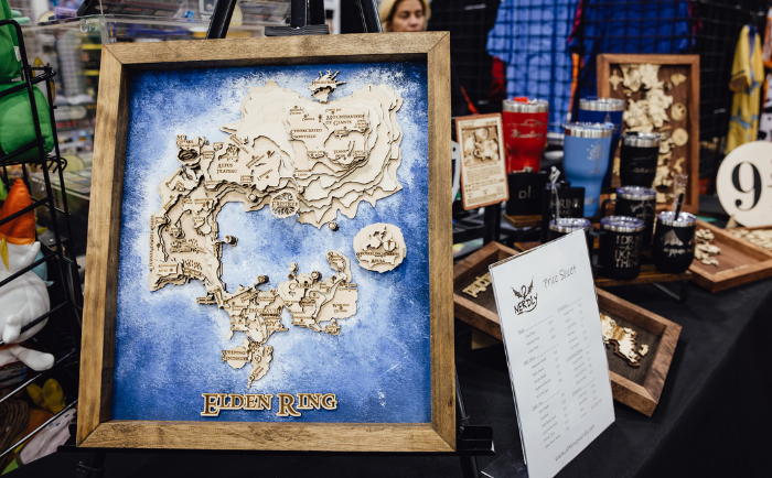 A framed 3D wooden map of the "Elden Ring" game world is showcased at a convention booth. The map features intricately carved geographical details against a blue watercolor-like background, with the title "ELDEN RING" prominently displayed at the bottom. Adjacent to the map, a price sheet labeled "Wood Nerds" is placed, suggesting the craft's maker or vendor. Surrounding the main display, other wooden crafts and merchandise can be seen, along with hints of attendees in the background, giving a sense of the bustling environment of the convention.