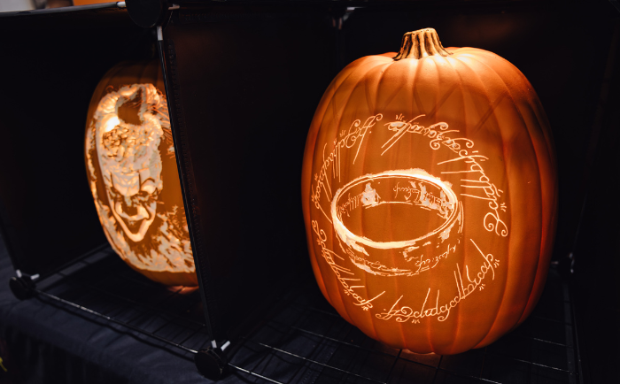 Two intricately carved pumpkins are illuminated from within, casting a warm glow that highlights their designs. The pumpkin on the left features a detailed face, while the one on the right showcases the iconic "One Ring" from "The Lord of the Rings", surrounded by its Elvish inscription. Both pumpkins are displayed on a black shelf, with the dark background further accentuating their luminance.