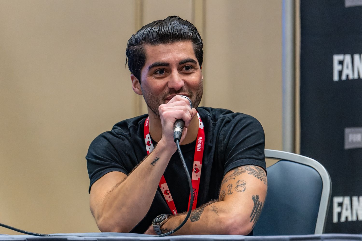 Hudson Thames sits at a table with a FAN EXPO mic in hand, smiling as he speaks to the audience. Two water bottles are set on the table in front of him.