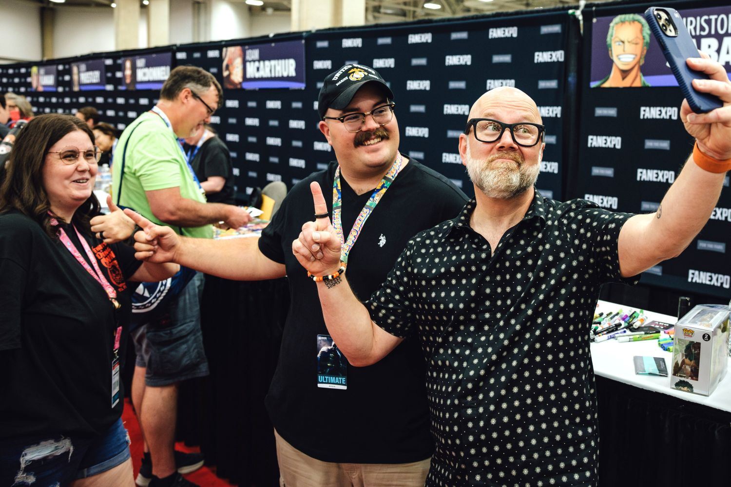 Voice actor Christopher Sabat poses for a selfie with fans during an autograph session at FAN EXPO. Everyone is smiling and pointing at the camera, with other attendees and voice actor booths visible in the background.
