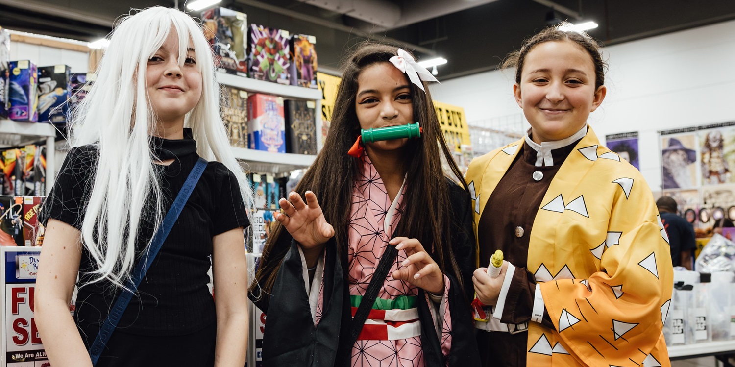 Three young fans in anime cosplay smile at the camera at Dallas FAN FESTIVAL. Costumes include characters from Demon Slayer, with colorful robes, a bamboo mouthpiece, and long white wig.
