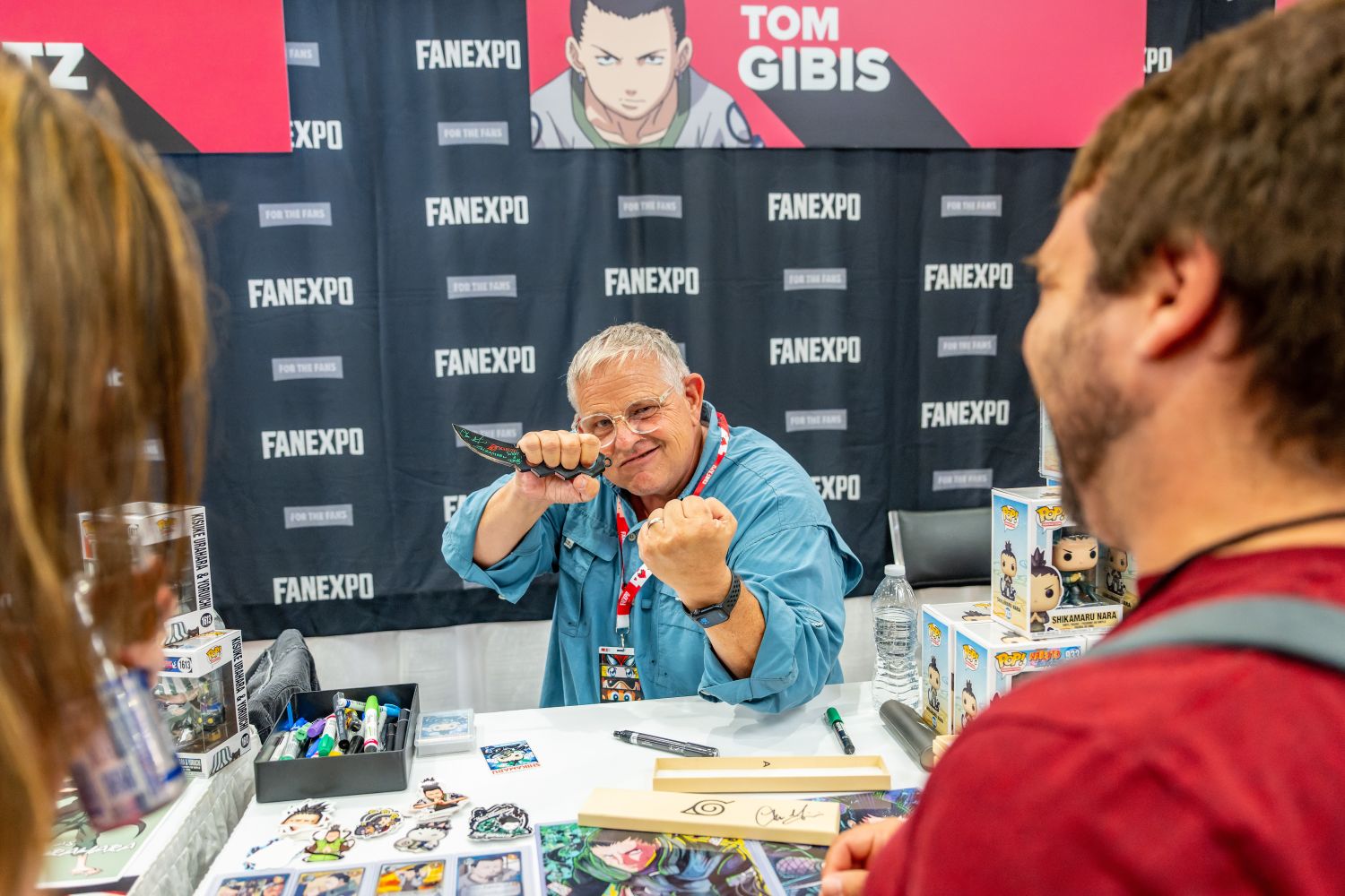 Voice actor Tom Gibis poses playfully at his table, raising his fists and holding a small prop kunai knife toward fans. His table is decorated with Naruto artwork and collectibles as attendees laugh and engage with him.