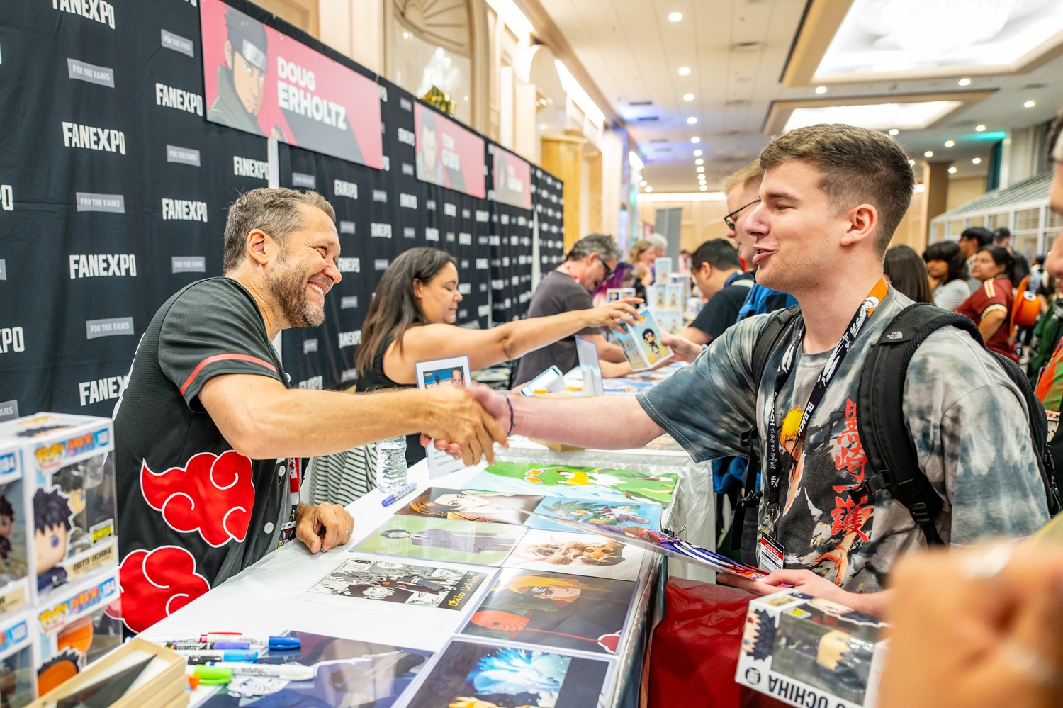 Voice actor Doug Erholtz shakes hands with a smiling fan across his autograph table. The table is covered with Naruto prints and Funko Pops, while other fans wait in line behind for their turn.