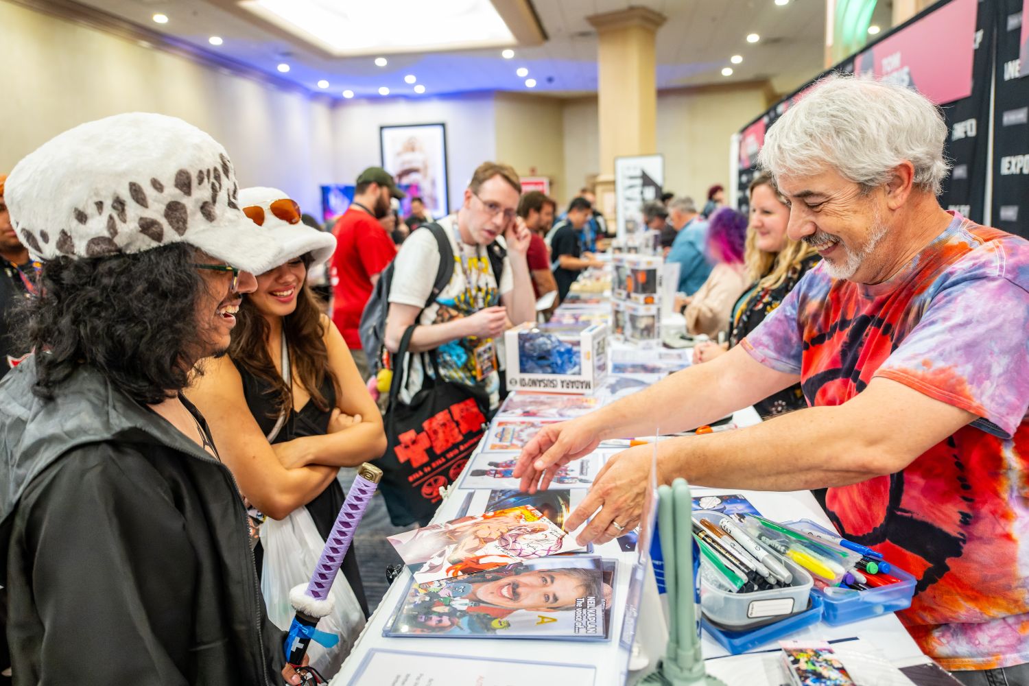 A guest artist in a tie-dye shirt smiles as he chats with two fans dressed in anime-inspired cosplay hats at his table. The table is covered with colorful prints, pens, and collectibles while the fans laugh and enjoy the interaction.