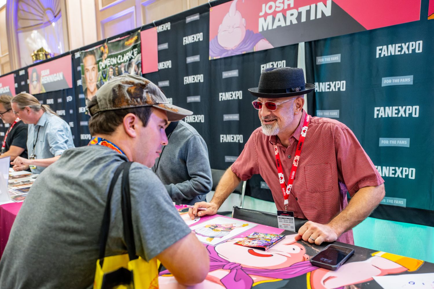 Voice actor Josh Martin, wearing a black hat and red sunglasses, leans forward across his table to chat warmly with a fan. The table is decorated with Dragon Ball Z art, including his character Majin Buu.