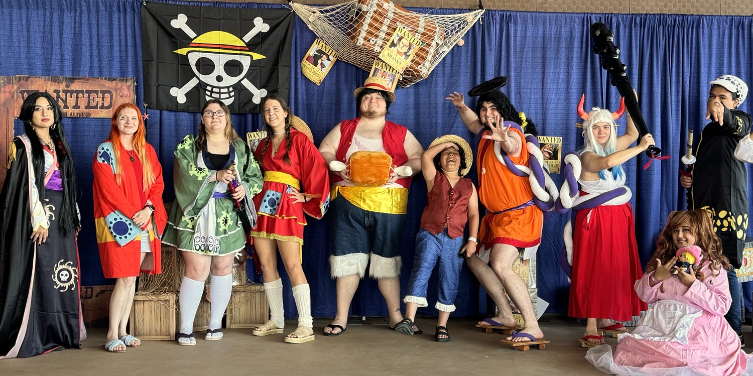 A large group of anime fans pose together in cosplay as characters from the popular series One Piece at an anime meetup during FAN EXPO Dallas. The group stands in front of a pirate-themed backdrop featuring a black flag with a straw hat skull-and-crossbones, wooden crates, and “WANTED” posters for various characters. Each cosplayer wears a unique, handmade costume representing characters like Luffy, Nami, Boa Hancock, and Yamato—complete with straw hats, props, and colorful wigs. One attendee holds a plush toy, while another wields a large black prop club. The group proudly smiles, strikes dynamic poses, and showcases the diversity and creativity of the anime cosplay community in a joyful, celebratory atmosphere.