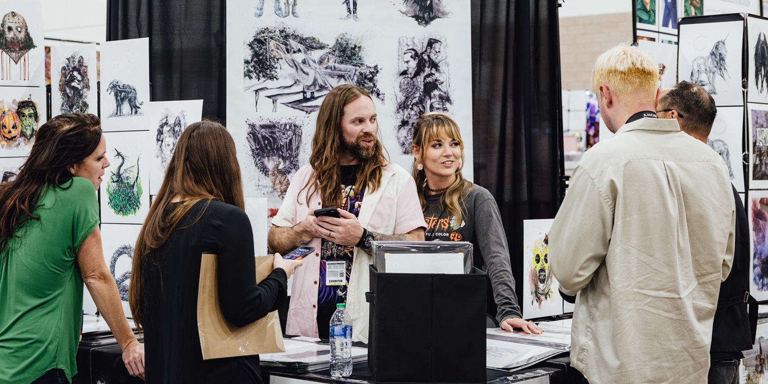 Artists chat with fans at their booth in Artist Alley at Dallas FAN FESTIVAL. Original artwork featuring fantasy creatures and horror-inspired designs covers the walls as attendees browse and commission pieces.