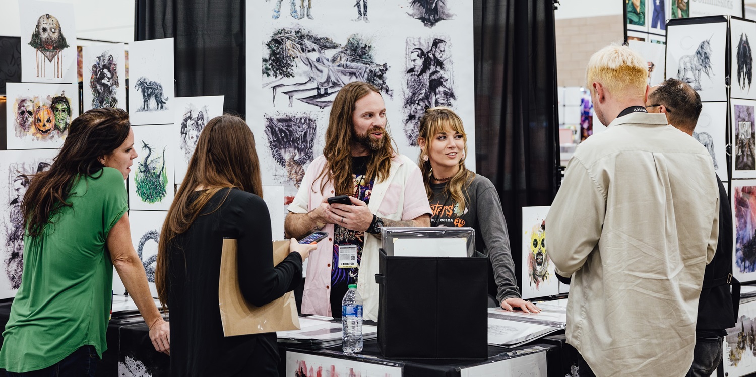 Artists engage with fans at a bustling booth in Artist Alley, showcasing original artwork. Various prints are displayed on the walls as attendees chat with the creators, making personal connections with the art community.