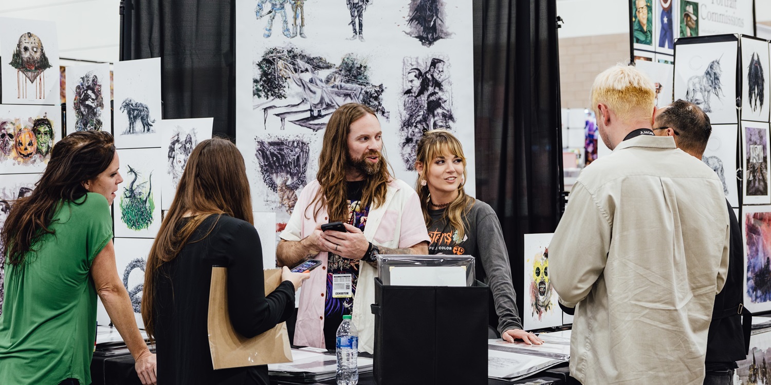 A group of attendees engage with an artist showcasing various horror-themed prints at a booth in Artist Alley during a convention.