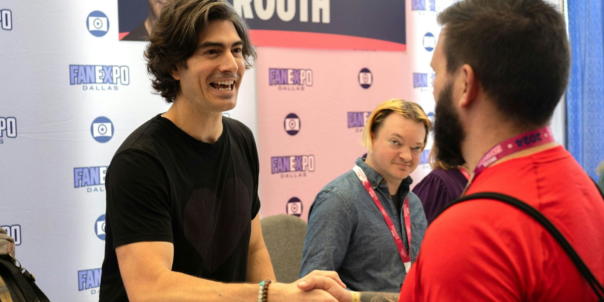 Actor Brandon Routh greets a fan with a handshake and a smile during an autograph session at FAN EXPO Dallas. A branded step-and-repeat backdrop with the FAN EXPO logo is visible behind him.