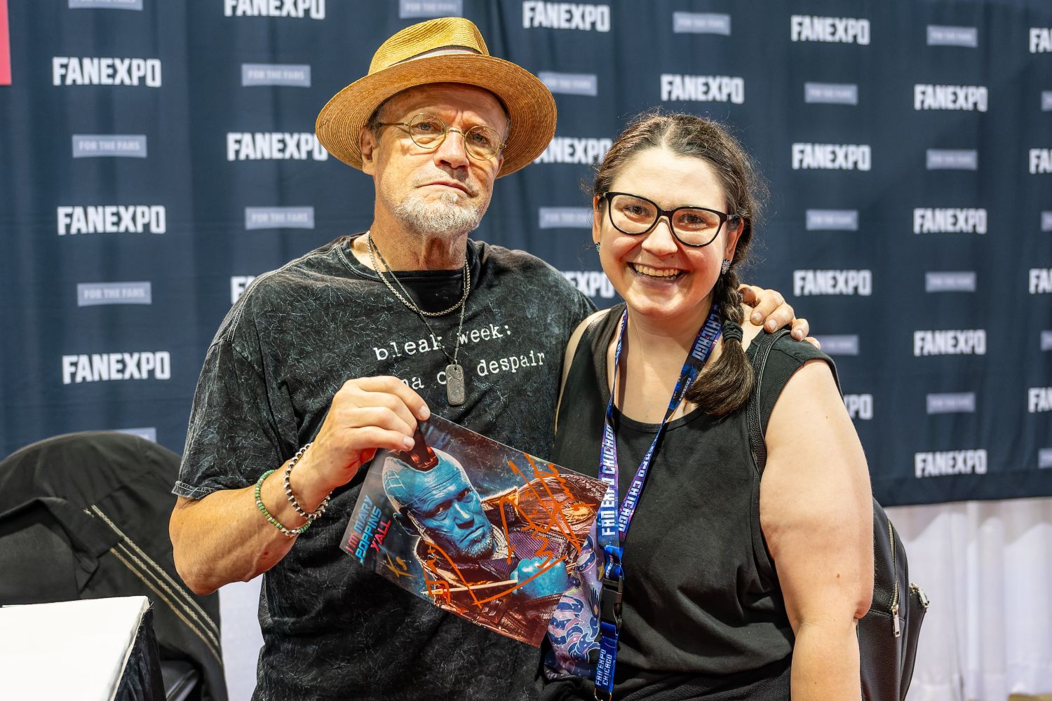 Michael Rooker stands beside a smiling fan in front of a FAN EXPO backdrop. He’s wearing a straw hat and dark t-shirt while holding up a signed print of his blue Yondu character from Guardians of the Galaxy. The fan grins wide, excited to be posing with him.