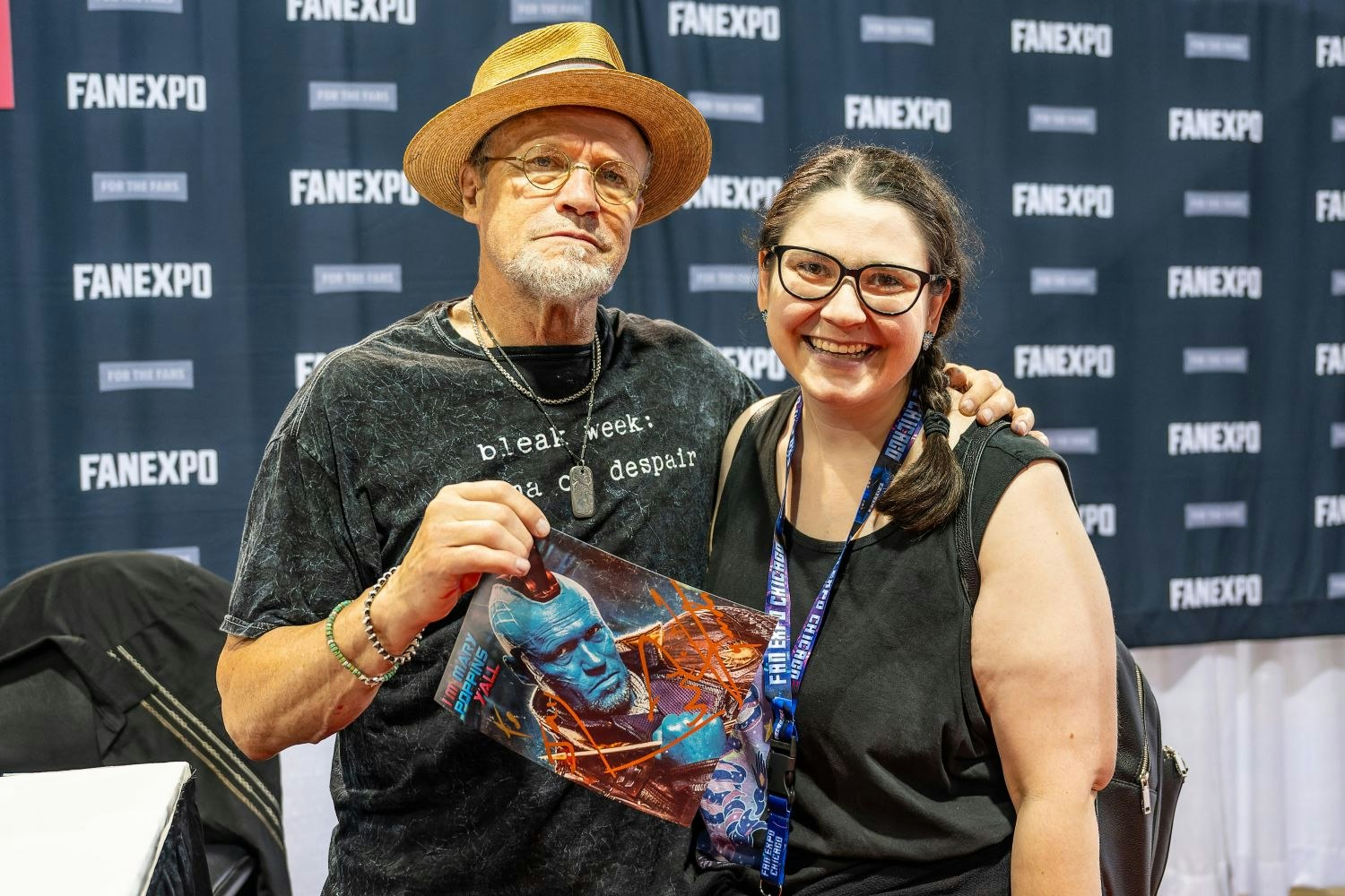 Michael Rooker stands beside a smiling fan in front of a FAN EXPO backdrop. He’s wearing a straw hat and dark t-shirt while holding up a signed print of his blue Yondu character from Guardians of the Galaxy. The fan grins wide, excited to be posing with him.