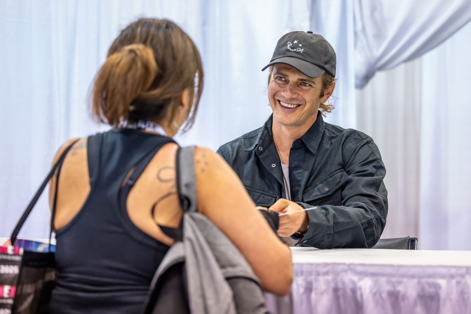 Hayden Christensen, dressed in a black jacket and baseball cap, beams warmly at a fan across the autograph table. The fan leans in with an item to be signed as Hayden listens and smiles, creating an approachable, genuine moment.