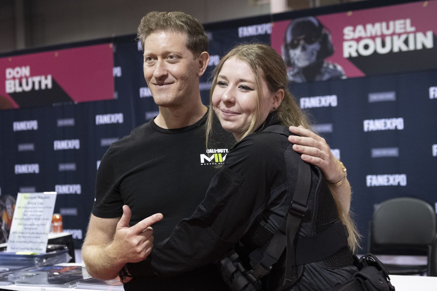 Samuel Roukin, in a black Call of Duty: Modern Warfare t-shirt, poses with a fan dressed in tactical cosplay gear. Standing in front of a FAN EXPO backdrop, he points playfully at the camera while the fan hugs him with a big smile.