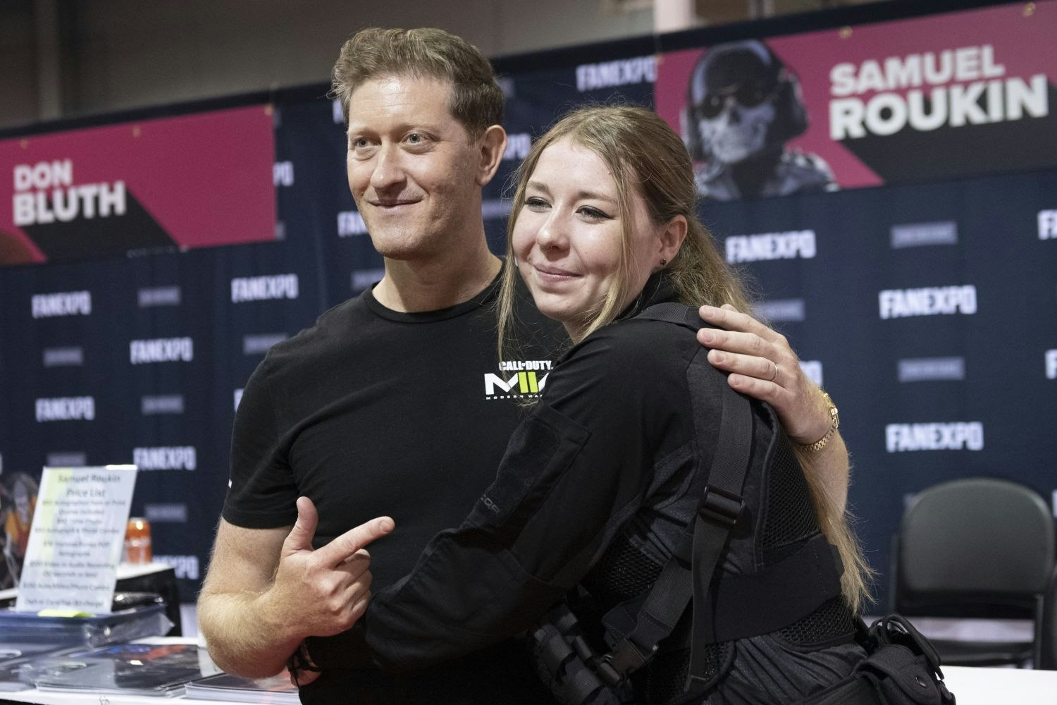 Samuel Roukin, in a black Call of Duty: Modern Warfare t-shirt, poses with a fan dressed in tactical cosplay gear. Standing in front of a FAN EXPO backdrop, he points playfully at the camera while the fan hugs him with a big smile.