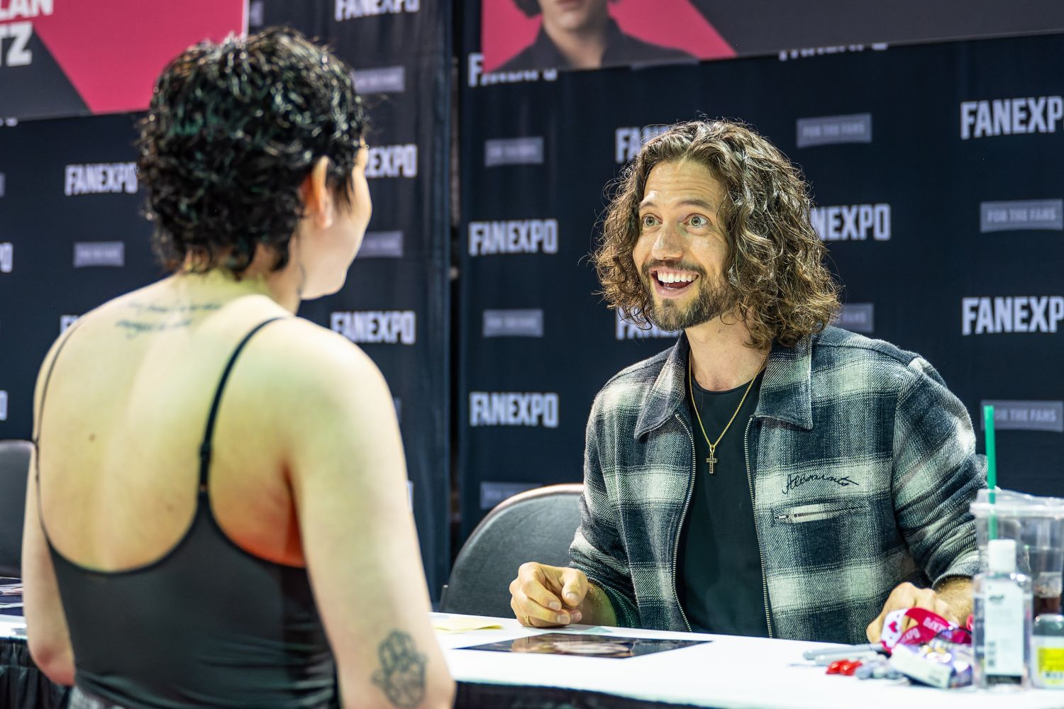 Jackson Rathbone sits behind his autograph table, looking thrilled as he chats with a fan. With his signature long curls and a plaid jacket, he smiles brightly, leaning in with excitement under a banner featuring his name and Twilight headshot.