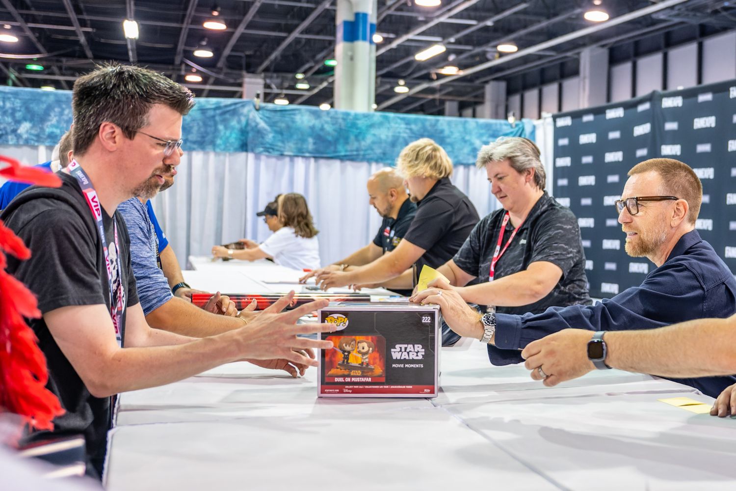 Ewan McGregor sits at a long autograph table alongside other guests, signing items for fans. In the foreground, a fan slides a boxed Star Wars Funko Pop across the table for him to sign, capturing a special moment between Obi-Wan and his admirers.